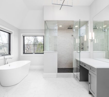 ELMHURST, IL, USA - APRIL 23, 2020: A large, new luxury bathroom with grey cabinets, white marble tile flooring, a freestanding bathtub with chrome faucet, and a glass walk-in shower.